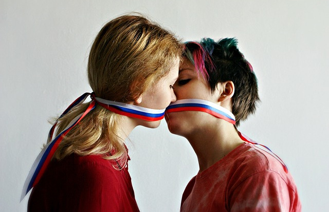 Thumbnail description followed by link description: Two white women kissing. The woman on the left is wearing a red top and has reddish hair. The woman on the right is wearing a pink top and has blue, pink, and brown hair. They are kissing with a white, blue, and red ribbon tied around their head's, covering their mouths.